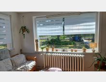 Helles Wohnzimmer mit großer Fensterfront und halb geöffneten Doppelrollos, breiter Fensterbank mit Pflanzen und Deko sowie einem Heizkörper darunter. Links steht ein Sofa neben einem Seitenfenster, rechts eine Stehlampe und Topfpflanze; durch das Fenster ist ein Gartenbereich sichtbar.