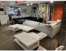 Ein Möbelgeschäft mit ausgestellten Sofas, Couchtischen und Teppichen. Im Hintergrund sind beleuchtete Wandlampen sowie stehende und hängende Teppiche in verschiedenen Farben und Mustern zu sehen.