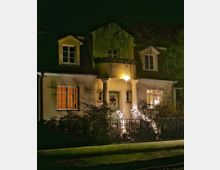 Einfamilienhaus bei Nacht mit leuchtender Weihnachtsdekoration, einschließlich eines beleuchteten Sterns über der Eingangstür und Lichterketten entlang des Treppengeländers, umgeben von einem kleinen Garten mit Zaun.