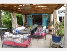 Überdachte Terrasse mit einer Holzkonstruktion und Backsteinpfeilern, möbliert mit schmiedeeisernen Sofas und gestreiften Kissen sowie einem langen Holztisch. Im Hintergrund sind leuchtend blaue Doppeltüren zu einem Innenraum geöffnet; umgeben von grünen Pflanzen und einem Garten.