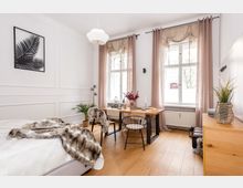 Helles Schlafzimmer mit Holzboden, einem Bett auf der linken Seite, das mit einer Felldecke bedeckt ist, und einem kleinen Essbereich mit Holztisch und schwarzen Stühlen. Große Fenster mit lichtdurchlässigen Vorhängen lassen viel Tageslicht herein, und Dekorationen wie Pflanzen und Kerzen schaffen eine wohnliche Atmosphäre.