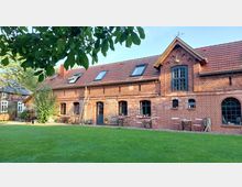 Backsteingebäude auf einem gepflegten Hof mit rotem Satteldach, mehreren Fenstern mit schwarzen Rahmen und kleinen Sitzgruppen vor der Fassade. Im Vordergrund eine große Rasenfläche, im Hintergrund weitere Fachwerkgebäude.