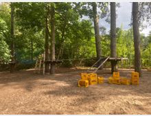 Ein Kletterwald mit Plattformen und Seilkonstruktionen, die zwischen hohen Bäumen aufgespannt sind, umgeben von dichtem Grün. Auf dem Boden stehen gelbe Getränkekisten, die anscheinend für Aktivitäten genutzt werden.