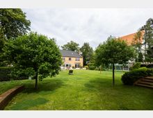 Großer Garten mit einer Rasenfläche, mehreren Bäumen und Büschen, im Hintergrund ein zweistöckiges Haus mit Backsteinfassade, schwarzem Dach und großen Fenstern. Auf der rechten Seite ist ein Spielgerüst und links eine gemauerte Treppe sichtbar.