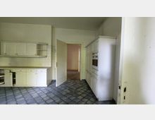 Helle Küche mit weißen Einbauschränken, Kacheln an den Wänden und dunkelgrauem, strukturiertem Bodenbelag. Eine geöffnete Tür führt zu einem angrenzenden Raum.