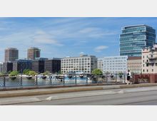 Moderne Stadtansicht mit Bürogebäuden, darunter ein Zalando-Gebäude, im Hintergrund; vorne eine Brücke mit dekorativem Geländer, die über einen Fluss führt, sowie eine Passantin auf dem Gehweg.