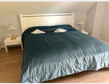 Schlafzimmer mit einem großen Bett mit weißem Kopfteil und blauer Bettdecke. Auf beiden Seiten stehen weiße Nachttische mit goldenen Lampen vor einer Wand mit blau-weiß gestreifter Tapete.