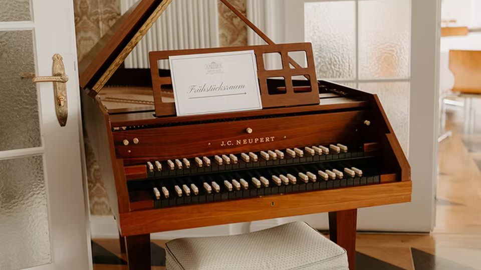 In einem hellen Innenraum steht ein hölzernes Tasteninstrument mit geöffnetem Deckel und gepolsterter Sitzbank auf einem schwarz-gemusterten Parkettboden. Im Hintergrund sind eine geriffelte Säule, gemusterte Tapete und weiße Glastüren zu einem angrenzenden Raum zu sehen.