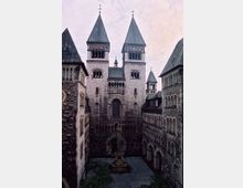 Romanische Kirche mit vier Türmen und einem kleinen Innenhof. Der Hof ist von steinernen Gebäuden mit Rundbogenfenstern umgeben und enthält einen zentralen Brunnen.