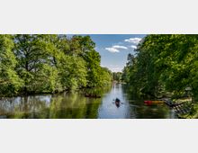 Ruhiger Fluss umgeben von dicht bewachsenen Bäumen, mit mehreren Kanus und Kajaks auf dem Wasser. Am rechten Ufer befinden sich ein Steg und eine kleine Anlegestelle.