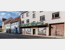 Straßenansicht mit alten Wohn- und Geschäftshäusern, darunter ein Gebäude mit verblasstem Schriftzug „Für Technik Haushalt“. Die Fassade zeigt historische Fotos in den Schaufenstern, daneben eine große Holztür mit diagonalem Muster.