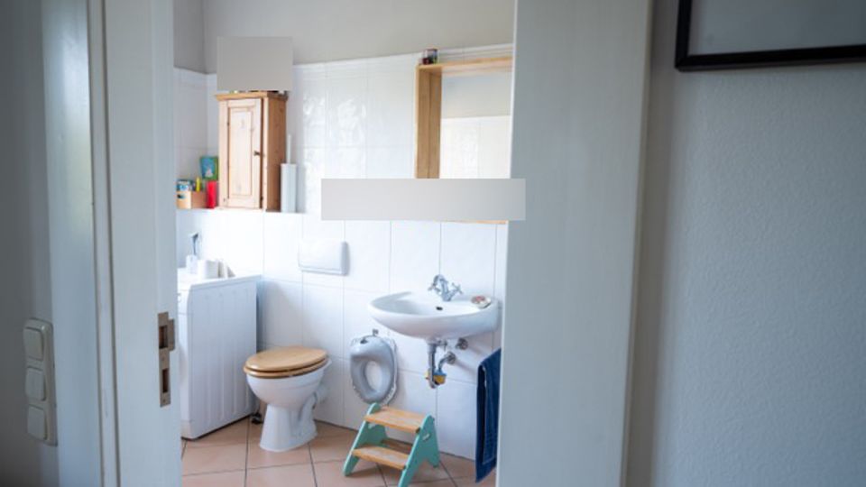 Blick durch eine Tür in ein kleines Badezimmer mit weißen Fliesen, rosa Bodenfliesen, WC und Waschbecken. An der Wand hängen ein Spiegel und ein kleiner Holzschrank, daneben steht ein Tritthocker und ein Heizkörper.