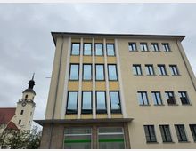 Mehrstöckiges modernes Gebäude mit cremefarbenen und gelben Fassadenelementen sowie großen Fenstern, daneben der Turm einer Kirche mit schwarzer Kuppel und Turmuhr. Im Vordergrund sind Bäume und im Erdgeschoss des Gebäudes ein Ladenschild sichtbar.