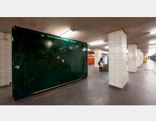 Eine U-Bahn-Station mit weiß gefliesten Säulen, einem dunkelgrünen Wandabschnitt mit Graffiti und einem Getränkeautomaten, vor dem eine Person steht. Der Boden ist aus dunklem Beton, und die Beleuchtung besteht aus rechteckigen Deckenlampen.