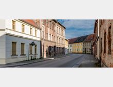 Straßenszene in einem historischen Stadtkern mit alten Wohnhäusern in verschiedenen Zuständen, teils renoviert und teils verfallen. Die Gebäude haben helle Fassaden und Ziegeldächer, die Straße ist leer und durch Bürgersteige gesäumt.