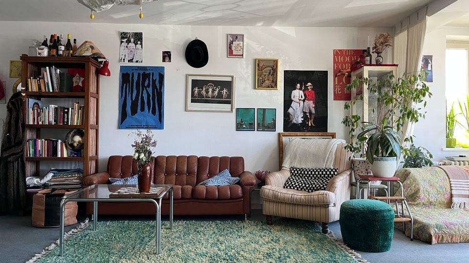 Cozy living room with a brown tufted sofa, a striped armchair, a glass coffee table on a large green rug, and a tall wooden bookcase. The white wall is decorated with framed art and posters, and large potted plants sit near a sunlit window on the right.