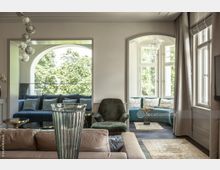 Helles Wohnzimmer mit modernen Möbeln, darunter ein beige Sofa, ein grüner Sessel und blau gepolsterte Sitzbänke vor großen Fenstern mit Blick auf Bäume. Ein Kronleuchter und Glasvasen ergänzen das minimalistische Design, durchsetzt von goldenen und blauen Akzenten.