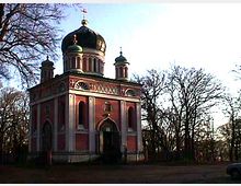 Orthodoxe Kirche mit rosa und weißen Fassaden, verziert mit Rundbögen und kleinen Türmchen. Das Gebäude hat eine große, goldene Kuppel und steht in einer von kahlen Bäumen umgebenen Landschaft.