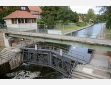 Schleusenanlage mit metallenen Schleusentoren und einer Brücke darüber, flankiert von Grünflächen und einem kleinen Gebäude mit rotem Ziegeldach; im Hintergrund ein Kanal, gesäumt von Bäumen und Wegen.