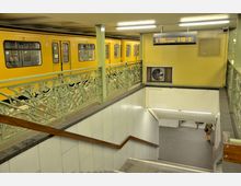 U-Bahn-Station mit gelbem Zug auf der oberen Ebene, verziertem grünen Geländer und einer Treppe, die zu einem unteren Bereich mit weiß gefliesten Wänden führt. An der Wand hängt eine digitale Anzeige mit Informationen zu den Abfahrtszeiten der Linie U7.