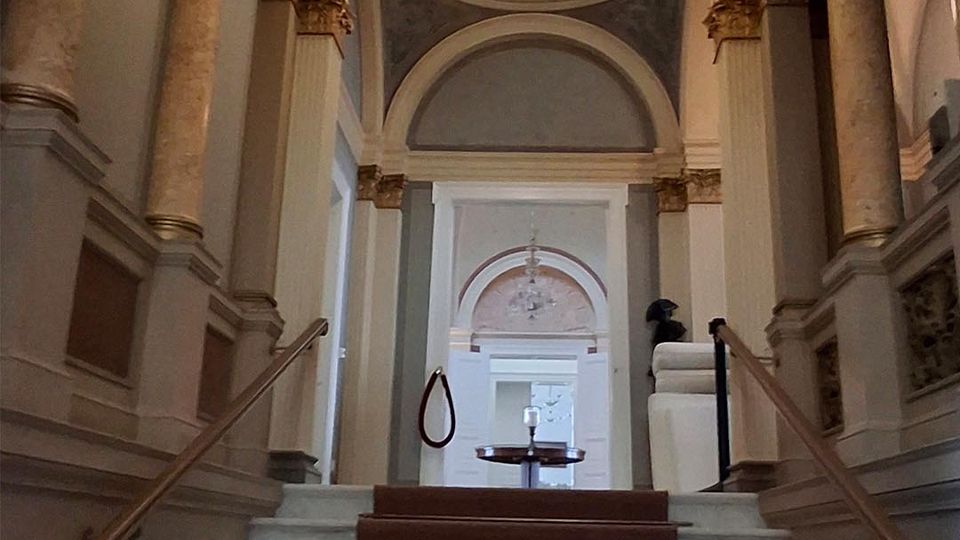 Blick in ein klassizistisches Treppenhaus mit breiter Marmortreppe und mittigem Teppichläufer, flankiert von Säulenreihen und Balustraden. Über den Treppen wölben sich hohe Rundbögen und eine Decke mit Oberlichtkuppel sowie hängender Leuchte.