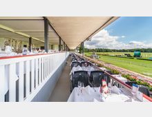 Überdachte Terrasse eines Rennbahngebäudes mit eingedeckten Tischen und schwarzen Stühlen. Im Hintergrund erstreckt sich eine grüne Rennbahn unter einem sonnigen Himmel mit einzelnen Wolken.