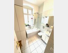 Helles, weiß gefliestes Badezimmer mit Badewanne und Glas-Duschabtrennung unter einem großen Fenster mit Rundbogen. Rechts befinden sich ein wandhängendes WC, ein Waschtisch mit Spiegel sowie ein hoher Schrank; der Boden ist mit großen quadratischen Fliesen ausgelegt.