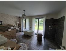 Großes, modernes Badezimmer mit freistehender Badewanne, ebenerdiger Dusche mit dunklen Wandfliesen und Holzfußboden. Eine Glas-Schiebetür mit transparenten Vorhängen führt auf eine Terrasse mit Blick auf einen Garten.