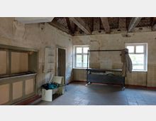 Ein verlassener, teils renovierungsbedürftiger Raum mit freiliegenden Holzbalken an der Decke, Putzrissen an den Wänden und zwei Fenstern, die Tageslicht hereinlassen. Im Raum befinden sich eine mobile Kleiderstange mit aufgehängten Jacken und Möbelstücke, darunter ein kleiner Schrank und eine Werkzeugkiste.