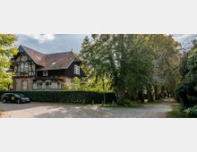 Fachwerkvilla mit rotem Ziegeldach, umgeben von einer Hecke und Bäumen, neben einer gepflasterten Straße und einem schattigen Weg durch eine Baumallee. Ein schwarzer Kombiwagen ist am Straßenrand geparkt.