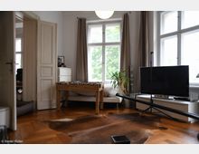 Wohnzimmer mit Parkettboden, einem großen Fenster mit beige Vorhängen, einem Fernseher auf einem Lowboard, einem Kickertisch und einer Zimmerpflanze. Im Vordergrund liegt eine braune, fellartige Teppichdeko.