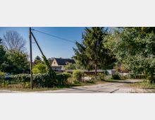 Ländliche Szene mit einem schmalen gepflasterten Weg, der an einem Zaun entlangführt, umgeben von Bäumen und Sträuchern. Im Hintergrund ist ein Wohnhaus mit dunklem Dach sowie kleine Unterstände sichtbar.