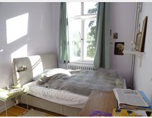 Schlafzimmer mit hellen, lavendelfarbenen Wänden, großem Fenster mit grünen Vorhängen und viel natürlichem Licht. Ein Bett mit grauer Decke steht neben einem kleinen grünen Beistelltisch mit Leselampe, und ein Holztisch mit Büchern befindet sich im Vordergrund.
