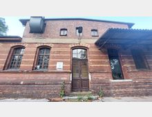 Backsteinfassade eines historischen Gebäudes mit hohen Rundbogenfenstern und einer verblassten hölzernen Doppeltür in der Mitte. An der Wand hängt ein Schild mit der Aufschrift „Haus 26“; rechts ist ein kleines Vordach aus Metall angebracht.