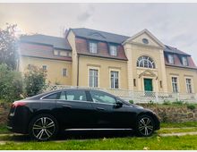 Ein historisches, zweistöckiges Gebäude mit beiger Fassade, rotem Ziegeldach und grüner Eingangstür. Im Vordergrund steht ein schwarzes Auto auf einer Grasfläche, umgeben von einem weißen Zaun und Garten.