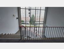 Treppenhaus mit großem Fenster, das den Blick auf einen gepflasterten Außenbereich und ein Hochhaus aus Backstein freigibt. Das Geländer ist schwarz mit einem roten Handlauf, und neben dem Fenster hängen geometrische Lampen von der Decke.