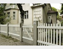 Beige Wohnhaus mit weißen Fensterrahmen, flankiert von einem Baum im Vorgarten. Ein niedriger, weißer Lattenzaun mit grauen Betonsäulen umgibt das Grundstück, der Gehweg davor ist mit Kopfsteinpflaster gestaltet.