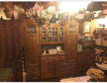 Traditionell eingerichtete Küche oder Wohnküche mit Holzmöbeln, darunter ein großer Holzschrank mit Glastüren, dekoriert mit Trockenblumen und Körben. Im Vordergrund steht ein Tisch mit einer Spitzen-Tischdecke und einem Blumenstrauß, die Beleuchtung kommt von einer rustikalen, hölzernen Deckenlampe.