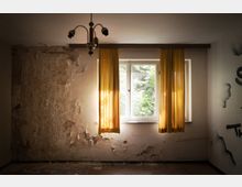 Ein verlassener Raum mit einer stark beschädigten Wand, abblätterndem Putz und einer alten Deckenlampe. Im Hintergrund ein einfaches Fenster mit gelben Vorhängen, durch das Tageslicht und Vegetation von außen sichtbar sind.