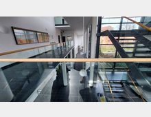 Ein modernes Gebäude mit einem Innenbereich, der ein Geländer aus Glas mit Holzhandlauf, eine Treppe mit Metall- und Glasdetails sowie große Fensterfronten zeigt, die viel Tageslicht einlassen. Im Hintergrund sind ein Gartenbereich und ein Backsteingebäude zu erkennen.