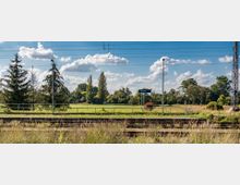 Bahnhofsumgebung mit Gleisen im Vordergrund und einer grünen Wiesenlandschaft im Hintergrund, umgeben von Bäumen. Ein Schild mit der Aufschrift „Zossen“ steht neben einer Laterne, unter einem wolkigen, blauen Himmel.