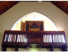 Holzempore in einer Kirche mit einer Orgel im Hintergrund, die hinter geöffneten, dekorativen Holztüren sichtbar ist. Die Wand dahinter ist in einem hellen Beige gehalten, und das Dach besteht aus dunklem Holz.