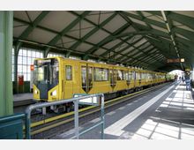 Eine gelbe U-Bahn steht an einem überdachten Bahnhof mit grüner Stahlkonstruktion und großen Fenstern. Menschen befinden sich auf dem Bahnsteig, und eine Anzeigetafel mit Informationen hängt von der Decke.