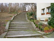 Steintreppe in einem Außenbereich, die durch eine leicht bewaldete Umgebung nach oben führt. Rechts befindet sich die Fassade eines Gebäudes mit zwei Fenstern, Blumenkästen und Efeubewuchs, daneben ein kleiner Garten mit Pflanzen und Sträuchern.