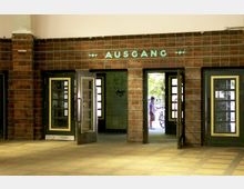 Backsteinwand mit grünen Türen und der Aufschrift „AUSGANG“ in türkisfarbenen Buchstaben. Durch eine geöffnete Tür ist der Blick auf einen Außenbereich mit Fahrrädern und einer Person zu sehen.