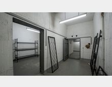 Ein leerer, betonierter Raum mit weißen Wänden und grauem Boden; im Vordergrund steht ein Metallrahmen an der Wand, während im angrenzenden Raum leere Metallregale stehen. Die Decke ist mit Neonlampen beleuchtet, und eine schwere Metalltür führt zu einem weiteren Bereich.