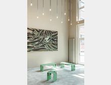 Ein minimalistischer, heller Raum mit hohen Wänden und großen Fenstern. An der Wand hängt ein abstraktes Kunstwerk mit wellenförmigen, grünen und dunklen Mustern, darunter stehen grüne, modulare Stühle und Tische auf einem grauen Betonboden, beleuchtet von hängenden, schlichten Lampen.