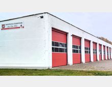 Weiße Garage der Freiwilligen Feuerwehr mit mehreren roten Rolltoren und einem Beschriftungsschild an der Wand. Der Bereich davor besteht aus gepflastertem Boden und einem schmalen Streifen mit Rasen.