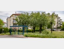 Bushaltestelle mit blauem Glasdach und Sitzbank, umgeben von Bäumen und Wiese. Im Hintergrund stehen mehrstöckige Wohngebäude mit Balkonen.