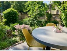 Runder Tisch mit gelber Sitzfläche und Vase mit rosa Blumen im Vordergrund, in einem grünen Garten mit blühenden Sträuchern und einer niedrigen Backsteinmauer im Hintergrund.